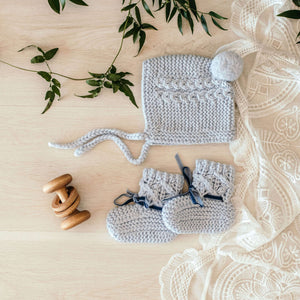 Blue merino wool knit bonnet and bootie set laying on a tablet with a wooden rattle