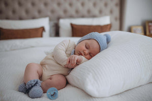 Baby laying on bed wearing a cream outfit and light blue bonnet and bootie set