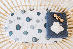 Rattan bassinet with cloud and hot air balloon sheet and folded blue blanket and  wooden rattle