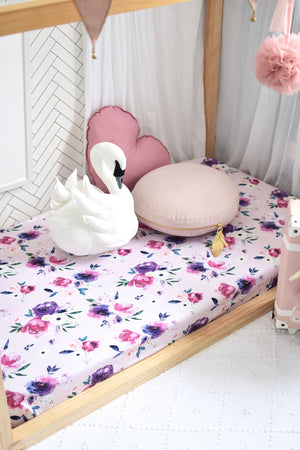 Pink and purple floral cot on wooden framed cot, with a white swan toy and nude round pillow and pink love heart pillow