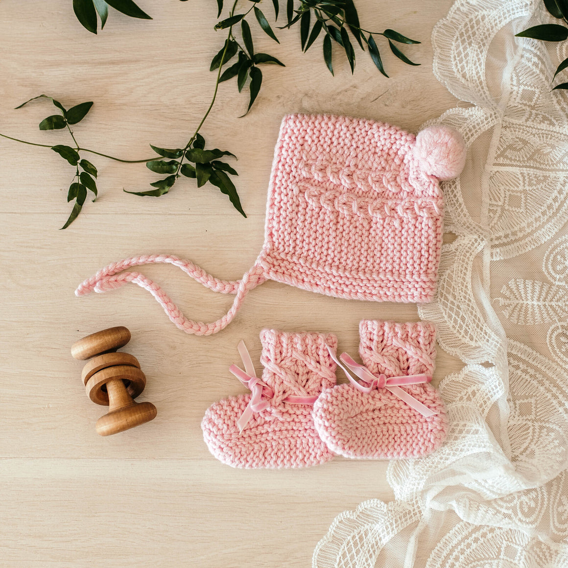 Pink merino wool bonnet and bootie set laying on a table and wooden rattle