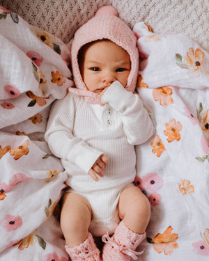 Baby girl laying on a floral swaddle wearing a white outfit and pink bonnet and bootie set