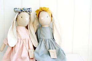 Alimorse Sofia Bunny wearing a pink dress and floral headband sitting with bunny wearing a blue dress and mustard headband