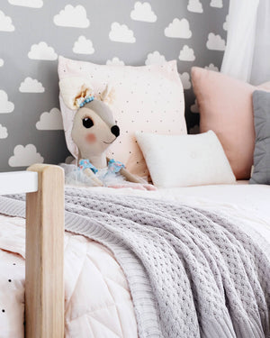 Grey knit blanket draped over white bed spread with teddy and pillows on it and grey and white cloud wall paper