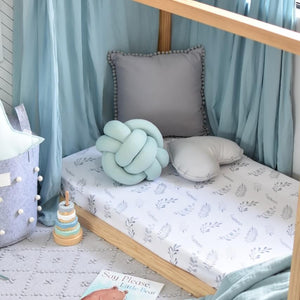 Toddler bed with white cot sheets which has ferns all over it, and has mint green canopy draped over toddler bed, grey basket sitting next to the bed and a stacker toy