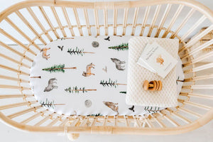 White bassinet sheet with pine trees, wolves and birds print on it, cream knit blanket and wooden rattle in bassinet