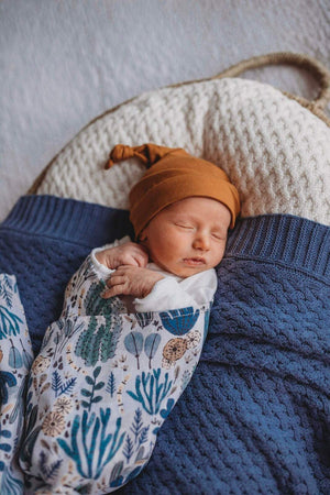 Newborn wrapped in a blue and white (arizona print) swaddle and mustard beanie, laying on a blue knit blanket