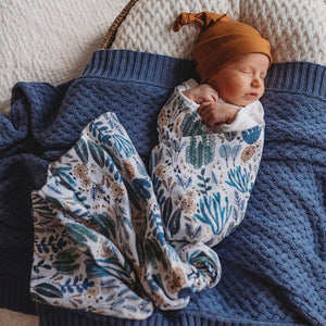 Newborn wrapped in a blue and white (arizona print) swaddle and mustard beanie, laying on a blue knit blanket