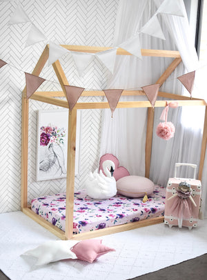 a wooden house framed cot with a pink and purple floral cot sheet, with a swan toy, pink heart pillow and nude round pillow, bunny artwork on the wall and bunting hanging from the ceiling