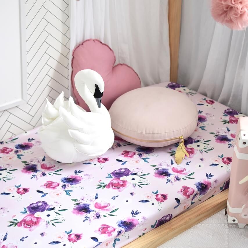 Pink and purple floral cot on wooden framed cot, with a white swan toy and nude round pillow and pink love heart pillow