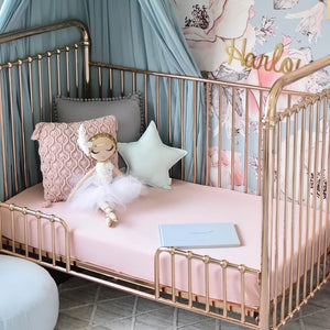Gold cot with pink cot sheets, three pillows and a doll sitting on cot, floral wall paper in background and green canopy