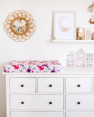 Photo of chest of drawers with pink and purple floral change pad cover, photos on the wall in the background and mirror on the wall