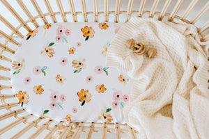 Rattan bassinet with a white sheet with pink and orange flower print and a cream knit blanket draped over the edge and wooden rattle