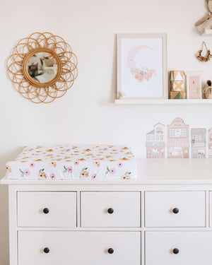 Chest of drawers with a white and pink and orange floral print change pad, photos and mirror on the wall in the background