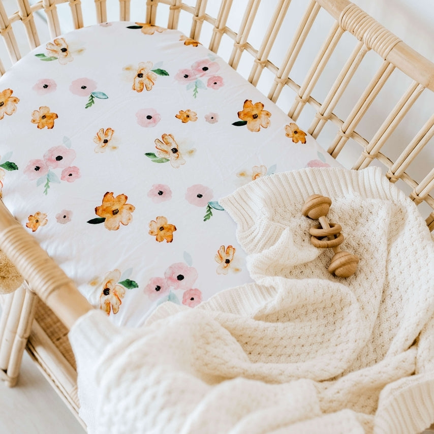 Rattan bassinet with a white sheet with pink and orange flower print and a cream knit blanket draped over the edge and wooden rattle