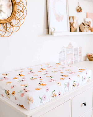 Chest of drawers with a white and pink and orange floral print change pad, photos and mirror on the wall in the background