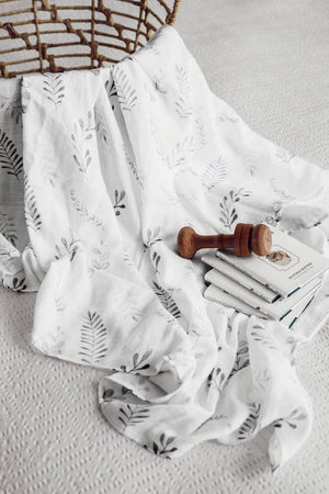 White swaddle with greens fern print draped over basket  and wooden rattle